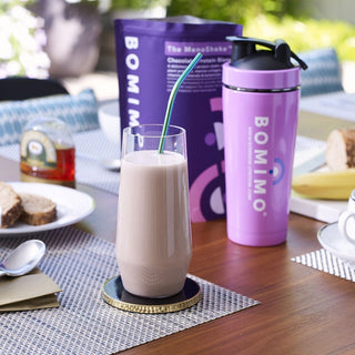 A glass of chocolate protein shake on a coaster in front of a packet of The Menoshake and the Bomimo protein shaker.