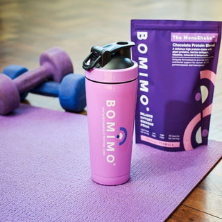 The Bomimo protein shaker in front of a packet of The Menoshake on a purple yoga mat with hand weights in the background.