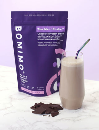 A packet of The Menoshake with a glass of  chocolate protein shake in front of it and  a small pile of chocolate next to it.
